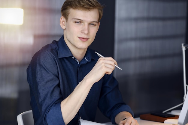 Jonge blonde zakenman die over strategie denkt op zijn werkplek met computer in een verduisterd kantoor, schittering van licht op de achtergrond.