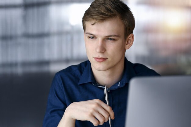 Jonge blonde zakenman die over strategie denkt op zijn werkplek met computer in een verduisterd kantoor, schittering van licht op de achtergrond.