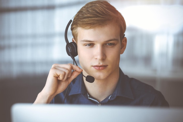 Jonge blonde zakenman met behulp van headset en computer in een verduisterd kantoor, schittering van licht op de achtergrond. opstarten betekent hard werken.