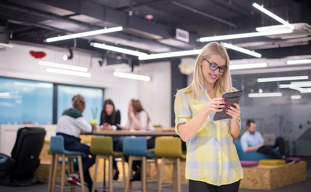 jonge blonde zakenvrouw die online werkt met behulp van digitale tablet terwijl ze op een modern startkantoor staat met een multi-etnisch zakelijk team op de achtergrond