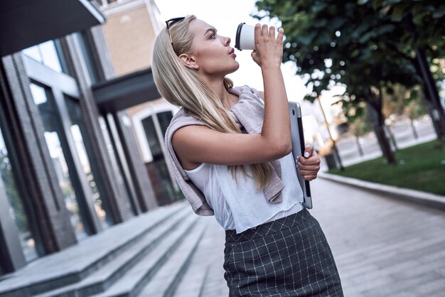 Jonge blonde zakenvrouw drinkt koffie en houdt laptop vast