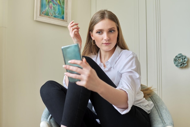 Jonge blonde zit thuis in de stoel met behulp van videocommunicatie op smartphone