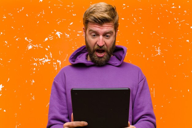 Jonge blondeman met een tablet die een paarse hoodie draagt tegen beschadigde oranje muur