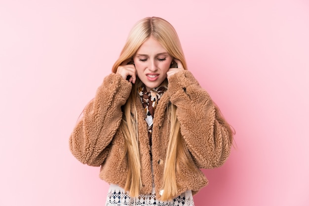 Jonge blondevrouw die een laag dragen tegen een roze muur die oren behandelen met handen.