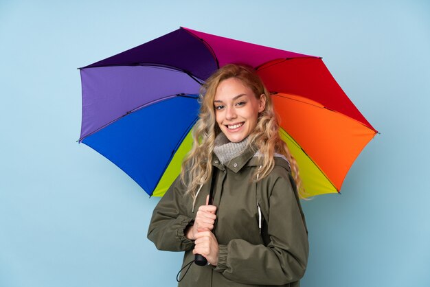 Jonge blondevrouw die een paraplu houden die op blauwe muur wordt geïsoleerd die veel glimlacht