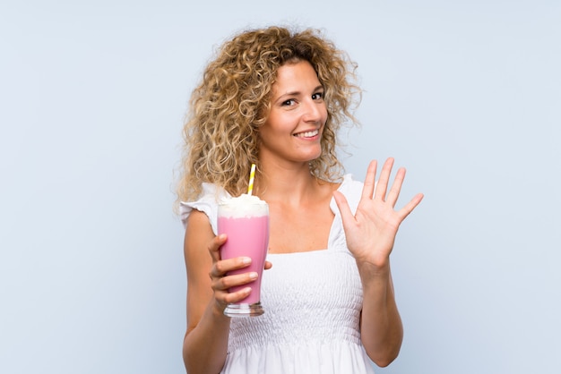 Jonge blondevrouw die met krullend haar een aardbeimilkshake groeten met hand met gelukkige uitdrukking houden