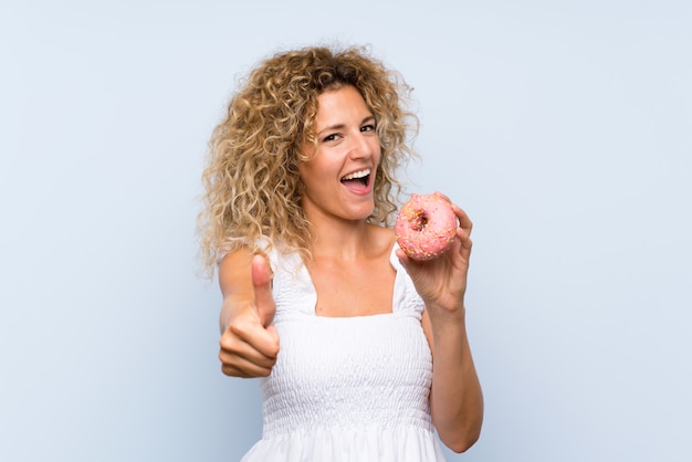 Jonge blondevrouw die met krullend haar een doughnut over geïsoleerde blauwe muur met omhoog duimen houden omdat iets goeds is gebeurd