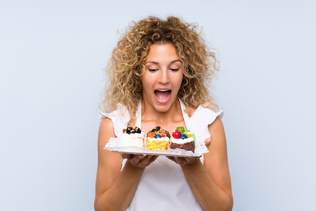 Foto jonge blondevrouw die met krullend haar veel verschillende minicakes houden