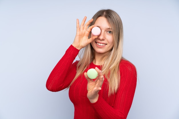 Jonge blondevrouw die over geïsoleerde blauwe muur kleurrijke Franse macarons houden