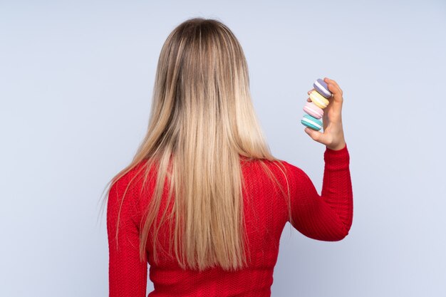 Jonge blondevrouw die over geïsoleerde muur kleurrijke Franse macarons in achterpositie houden