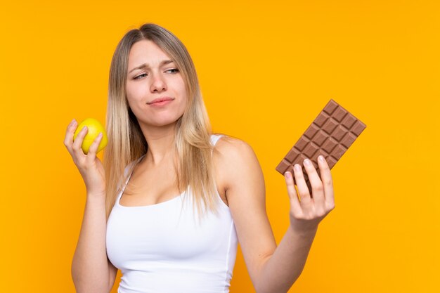 Jonge blondevrouw die twijfels hebben terwijl het nemen van een chocoladetablet in één hand en een appel in de andere