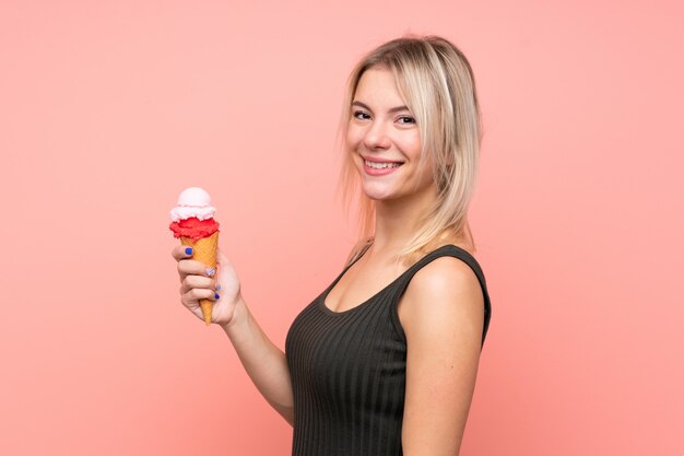 Jonge blondevrouw met een cornetroomijs over geïsoleerde roze muur die veel glimlacht