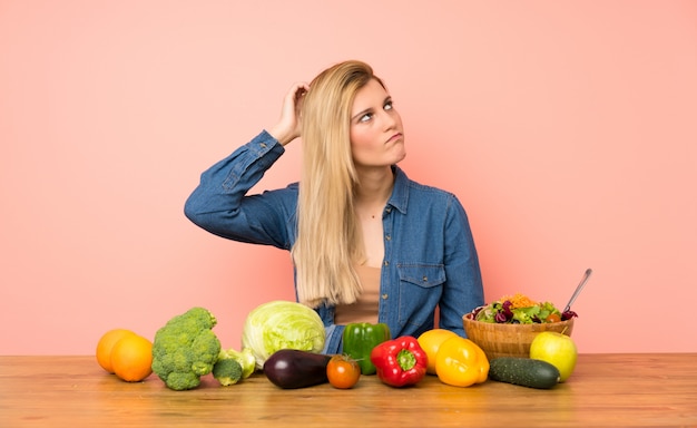 Jonge blondevrouw met vele groenten die twijfels hebben terwijl het krassen van hoofd