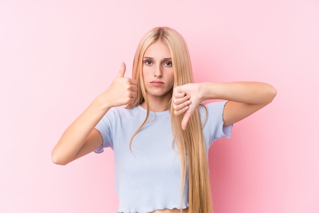 Jonge blondevrouw op roze muur die duimen tonen en duimen neer, moeilijk concept