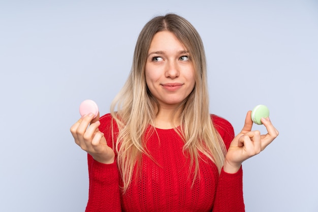 Jonge blondevrouw over blauw die kleurrijke Franse macarons en het denken houden