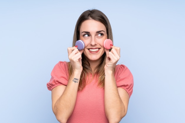 Jonge blondevrouw over geïsoleerde blauwe kleurrijke macarons houden en muur die omhoog kijken