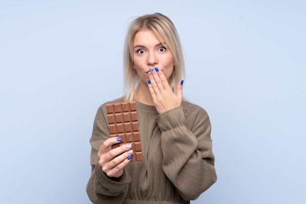 Jonge blondevrouw over geïsoleerde blauwe muur die een chocoladetablet nemen en verrast