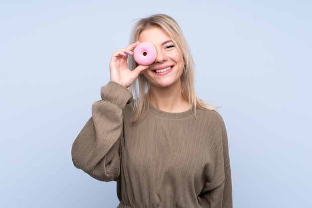 Jonge blondevrouw over geïsoleerde blauwe muur die een doughnut houden en gelukkig