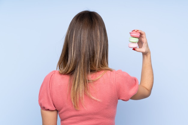 Jonge blondevrouw over geïsoleerde blauwe muur die kleurrijke Franse macarons in achterpositie houden