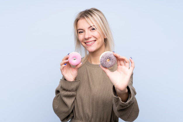 Jonge blondevrouw over geïsoleerde blauwe muurholding donuts met gelukkige uitdrukking