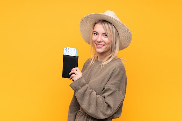 Jonge blondevrouw over geïsoleerde gele muur gelukkig in vakantie met paspoort en vliegtuigtickets