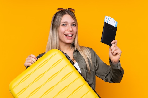 Jonge blondevrouw over geïsoleerde gele muur in vakantie met koffer en paspoort