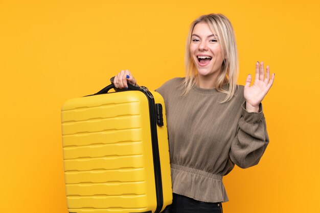 Jonge blondevrouw over geïsoleerde gele muur in vakantie met reiskoffer en het maken van OK teken