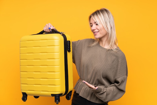 Jonge blondevrouw over geïsoleerde gele muur in vakantie met reiskoffer