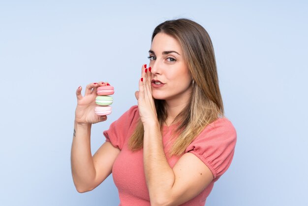 Jonge blondevrouw over geïsoleerde muur die kleurrijke Franse macarons houden en iets fluisteren