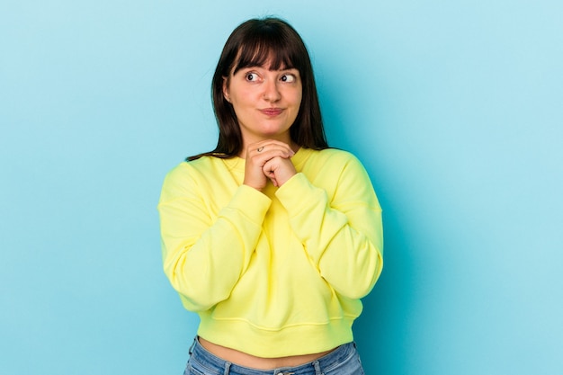 Jonge bochtige blanke vrouw geïsoleerd op blauwe achtergrond houdt handen onder de kin, kijkt gelukkig opzij.