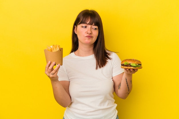 Jonge bochtige blanke vrouw met een hamburger geïsoleerd op gele achtergrond