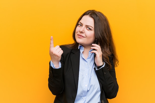 Jonge bochtige plus size zakenvrouw houden een telefoon wijzend met vinger naar je alsof uitnodigen dichterbij komen.