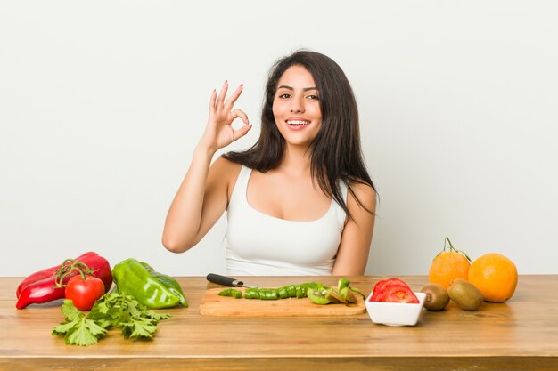 Jonge bochtige vrouw die een gezonde maaltijd voorbereidt vrolijk en zeker tonend ok gebaar.