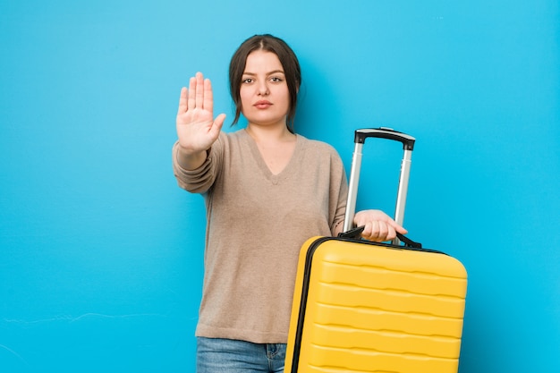 Jonge bochtige vrouw die een koffer houdt die zich met uitgestrekte hand bevindt die eindeteken toont, dat u verhindert.