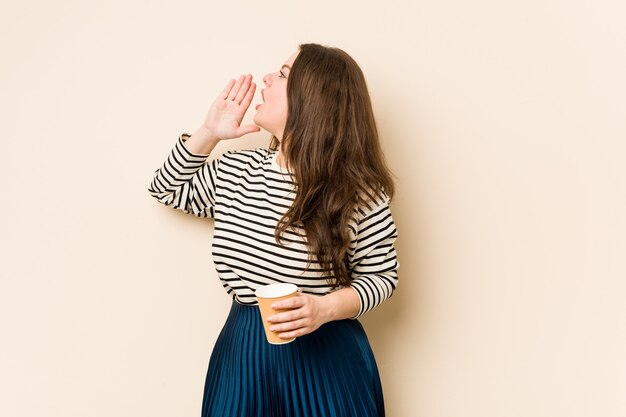 Jonge bochtige vrouw die een koffie houdt die en palm dichtbij geopende mond houdt houdt.