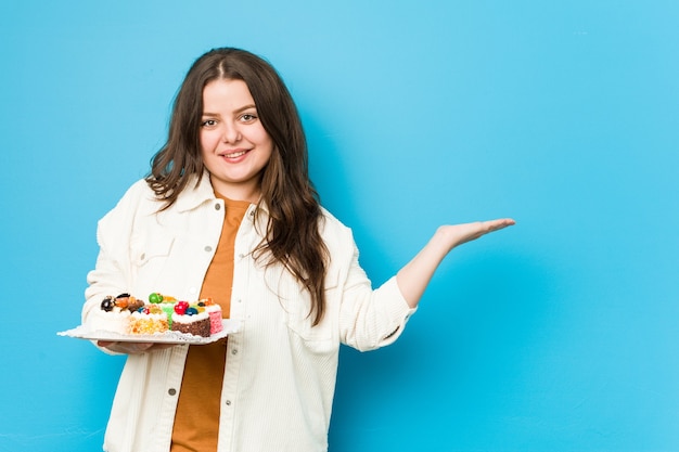 Jonge bochtige vrouw die zoete cakes houdt die een exemplaarruimte op een palm tonen en een andere hand op taille houden.