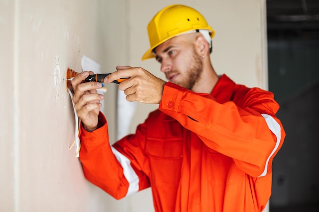 Jonge bouwer in oranje werkkleding en gele veiligheidshelm die bedachtzaam een schroevendraaier gebruikt op het werk in de flat