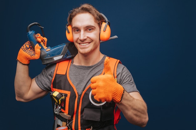 Jonge bouwer vertoont duim terwijl hij apparatuur in de arm heeft