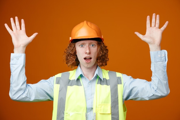 Foto jonge bouwvakker in bouwuniform en veiligheidshelm die naar de camera kijkt, verrast zijn armen in overgave op te heffen over oranje achtergrond