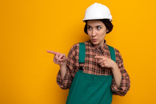 Jonge bouwvrouw in bouwuniform en veiligheidshelm die opzij kijkt met een serieus gezicht wijzend met wijsvingers naar de zijkant die op oranje staat