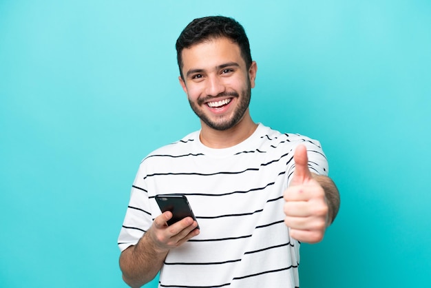 Jonge Braziliaanse man geïsoleerd op blauwe achtergrond met behulp van mobiele telefoon terwijl het doen van thumbs up