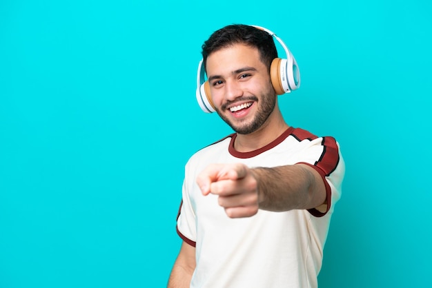 Jonge Braziliaanse man geïsoleerd op een blauwe achtergrond die muziek luistert en naar voren wijst