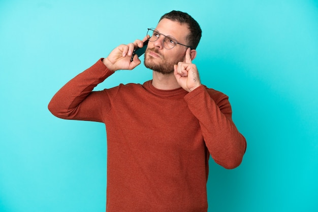 Jonge Braziliaanse man met behulp van mobiele telefoon geïsoleerd op blauwe achtergrond met twijfels en denken