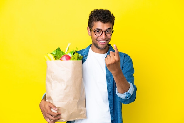 Jonge Braziliaanse man met een boodschappentas geïsoleerd op een gele achtergrond die een komend gebaar doet