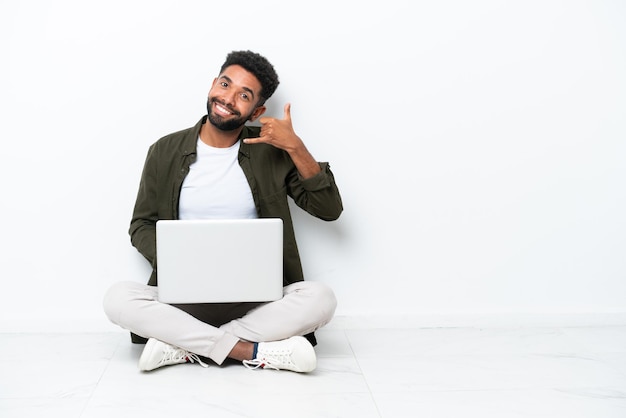 Jonge Braziliaanse man met een laptop zittend op de vloer geïsoleerd op wit telefoongebaar maken Bel me terug teken