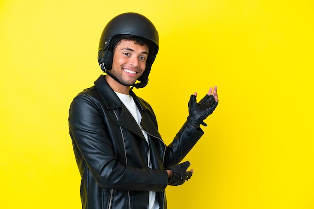 Jonge Braziliaanse man met een motorhelm geïsoleerd op een gele achtergrond die zijn handen naar de zijkant uitstrekt om uit te nodigen om te komen