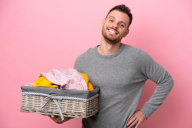 Jonge Braziliaanse man met een wasmand geïsoleerd op roze achtergrond poseren met armen op heup en glimlachend