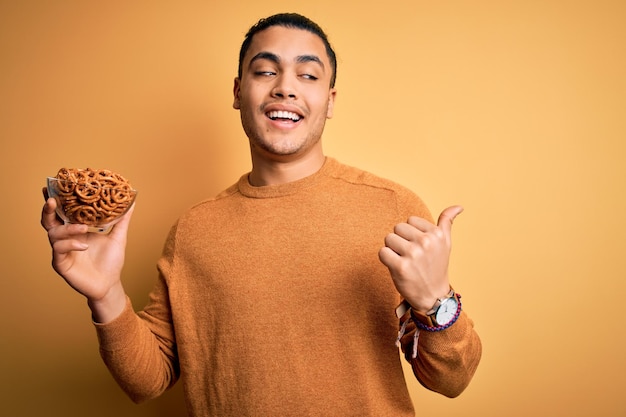 Jonge braziliaanse man met kom met duitse zoute gebakken pretzels over gele achtergrond wijzend en tonend met duim omhoog naar de zijkant met blij gezicht lachend