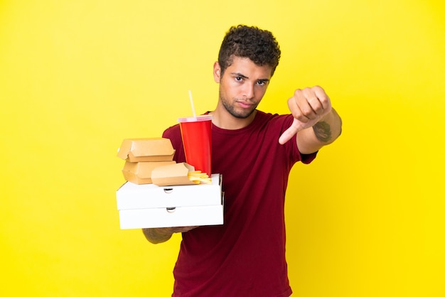 Jonge braziliaanse man met pizza's en hamburgers geïsoleerde achtergrond met duim omlaag met negatieve uitdrukking