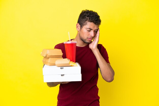 Jonge Braziliaanse man met pizza's en hamburgers geïsoleerde achtergrond met hoofdpijn